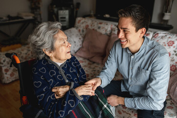 Young man sitting next to an old sick aged woman in wheelchair taking her hands while talking and smiling..