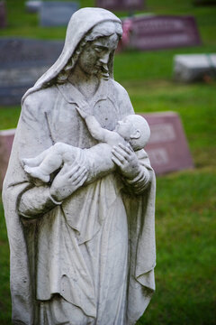 Mother cradling dead baby stone cemetery statue. Somber image with gravestones in the defocused background