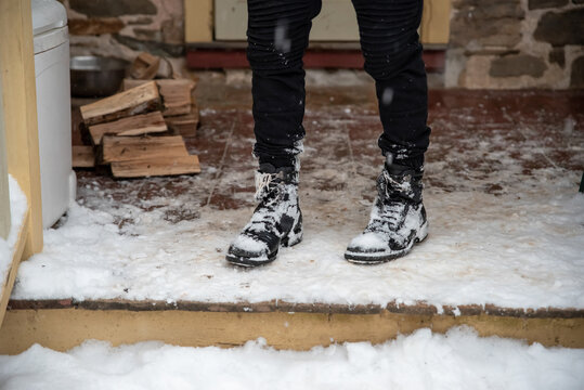 Bringing in firewood on a snowy day. Snowy boots and legs on a porch by a stack of firewood. Winter image with snow on the ground.