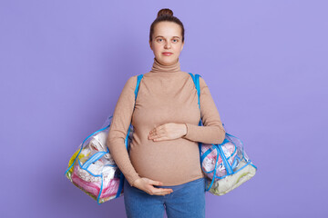 Young attractive pregnant woman wearing casual attire holding two bags with mother and child stuff, touching her belly with hands, looks at camera, being about going to maternity house.