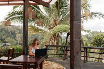 Girl freelancer work typing on laptop and looking at monitor with beautiful view, palm trees. . Traveling with a computer. Online dream job on the island