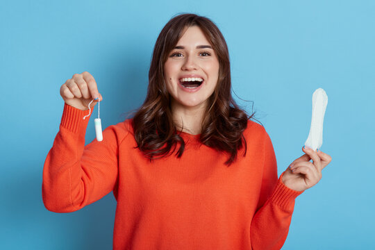 Young attractive woman holds cotton tampon and sanitary napkin, has excited look, monthly menstrual cycle. Protective care for women health. Girl with swab and pad against blue background.