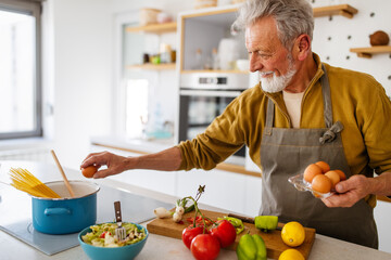 Happy retired senior man cooking in kitchen. Retirement, hobby people concept - obrazy, fototapety, plakaty