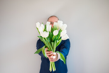 The man in blue shirt is standing with white tulips hiding his face on gray background. Place for text. 