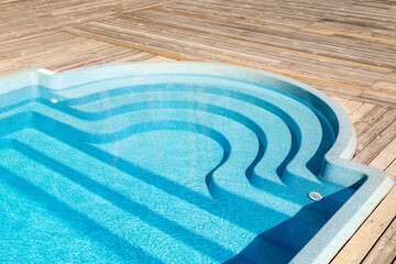 New modern fiberglass plastic swimming pool entrance step with clean fresh refreshing blue water on bright hot summer day at yard or resort hotel spa area. Wooden flooring deck of teak or larch board