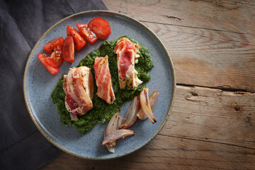 Fried  chicken breast fillet rolled in bacon on cream spinach with roasted onions and tomatoes on a blue plate and a rustic wooden table, copy space, high angle view from above
