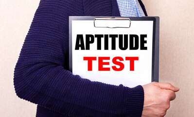 APTITUDE TEST is written on a white sheet held by a man standing sideways