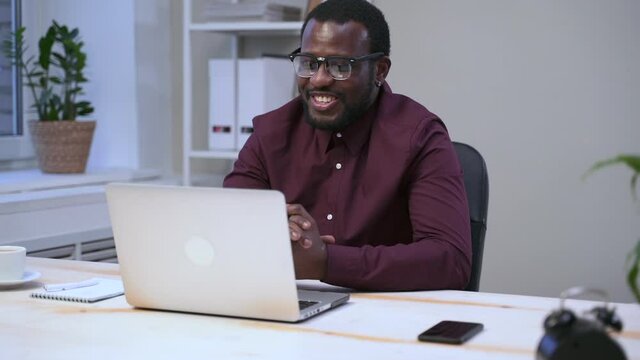 Young businessman communicates and uses laptop while sitting at table in modern office spbas. American man talking with happy smile and looking at computer screen during online conference at desk in
