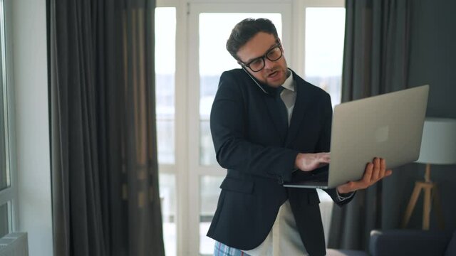 Emotional caucasian man with glasses dressed in a jacket and underwear negotiates using smartphone and laptop at home in a modern apartment