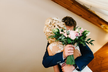 The groom in suit hugs bride in white dress are holding a beautiful wedding bouquet at home. Portrait of a beautiful young couple.