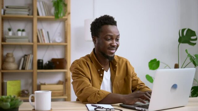 Man using laptop computer and having fun, sitting at table in home room spbas. Young guy looks at screen of device with smile and does remote job, sits in light workplace. American African person uses