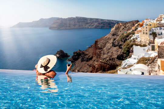 A woman with hat and a drink in her hand enjoys the view from an infinty swimming pool to the beautiful village of Oia on Santorini island, Greece