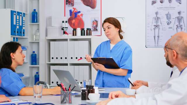 Team Of Doctors Discussing Diagnosis Sitting At Table In Clinic, Nurse Holding Clipboard Sharing Tasks To Colleagues. Group Of Physicians Talking About Symptoms Of Disease In Clinic Office