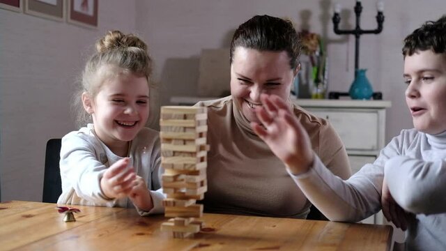 Happy young adult mom playing with two cute kids. A playful mom helps preschool children build a tower of wooden blocks in the living room. Educational games with the whole family.