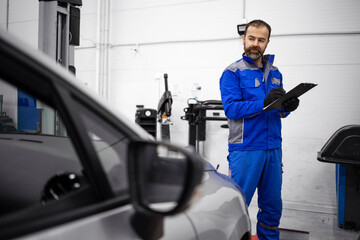 Middle aged bearded caucasian car mechanic doing visual inspection of vehicle in workshop and holding checklist.