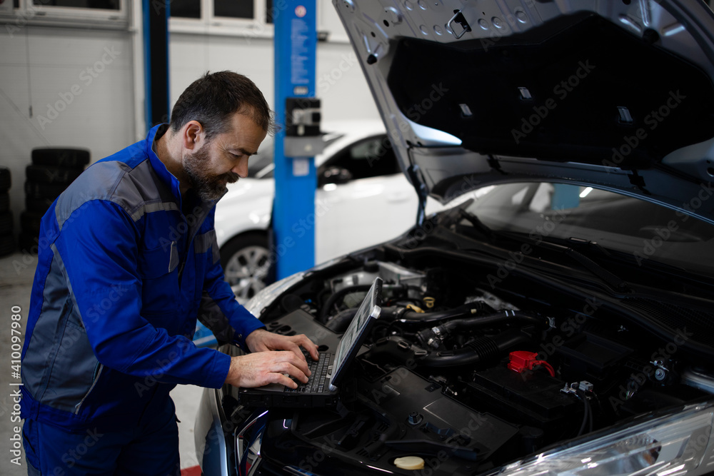 Wall mural Professional middle aged caucasian car mechanic with laptop computer diagnostic tool standing by vehicle engine area with hood open detecting malfunction.