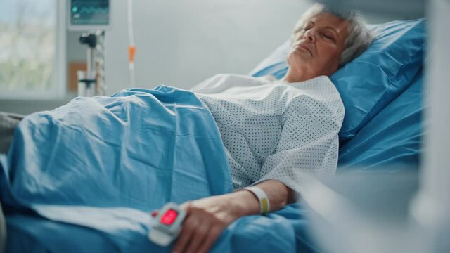 Hospital Ward: Senior Woman Resting in a bed with Finger Heart Rate Monitor / Pulse oximeter showing Pulse. Her Fragile Hands Resting on a Blanket. Focus on the Hand. Deeply Emotional Cinematic Shot