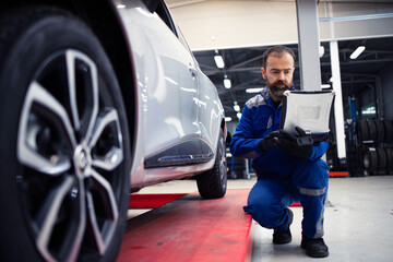 Professional middle aged bearded caucasian car mechanic doing visual inspection of vehicle in workshop and diagnostic tool. Automobile service and maintenance.