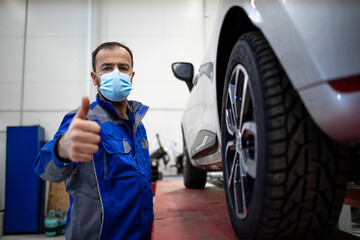 Fototapeta na wymiar Portrait of professional car mechanic wearing face mask due to corona virus standing in vehicle workshop by an automobile.