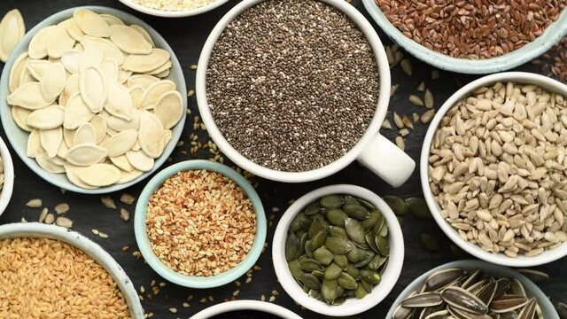  Assortment od healthy seeds - sesame, flax seed, sunflower seeds, pumpkin seed, chia and black seed in bowls on a black stone background. Top view