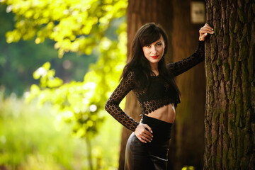 sexy brunette girl in a lace blouse and leather skirt with high heels posing near a tree in the...