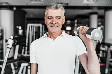 Fototapeta na wymiar Close up of a smiling senior man in the middle of the gym with a dumbbell in his hands. Training, health
