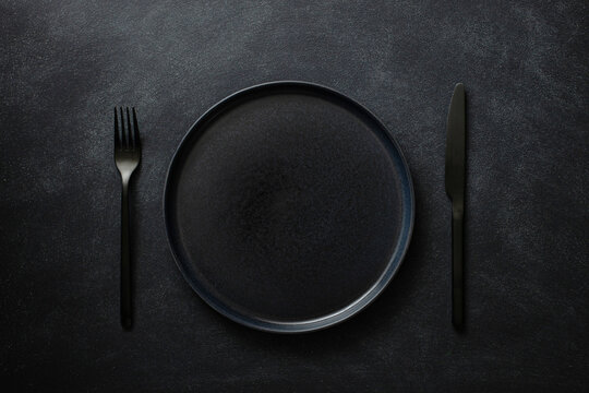 Empty black plate with black cutlery on the black table. Top view.