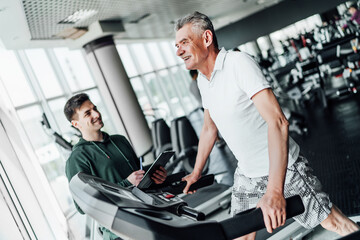 Fototapeta na wymiar The senior man on the treadmill is smiling and full of energy. His tenor is standing next to him and watching him