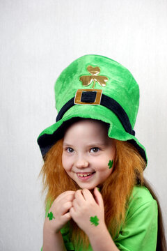 Laughting cute small girl with decorative red beard, green shamrok leaf on her cheek and leprechaun hat, raised her hands with green nails and clover leaves