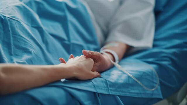 Hospital Ward: Senior Woman Resting and Recovering in Bed, Caring Daughter Holds Her Fragile Hand in Gesture of Support and Comforting. Focus on the Hands. Deeply Emotional Cinematic Shot