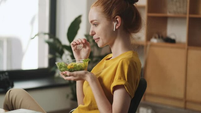 A focused young redhead woman with earbuds is eating salad while watching laptop sitting in the office