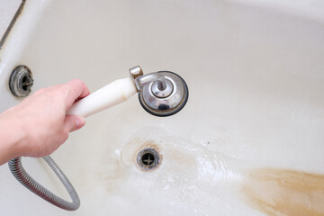 A hand holding shower head with running water against rusty bathtube with limescale, bathroom clining and decalcifying