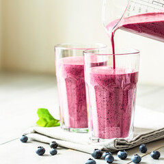 Two glasses of blueberry smoothie with mint garnish and straw on the table. Berry shake is poured...