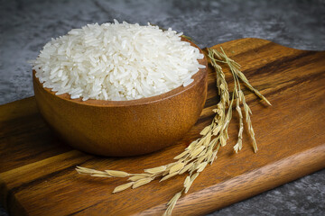 Jasmine rice in a wooden bowl placed on a dark wooden cutting board with rice stalks, rice and...