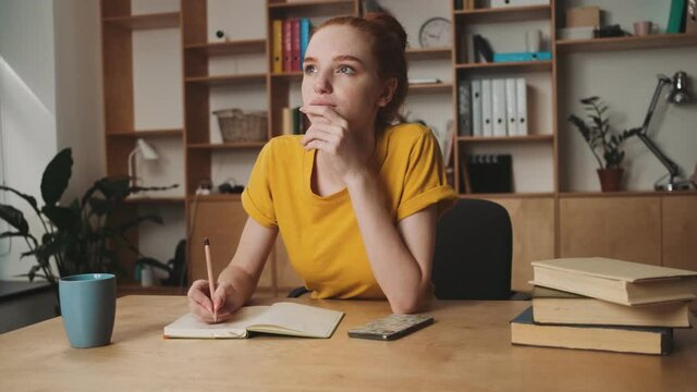 A thinking young redhead woman is writing something sitting at the table in the office