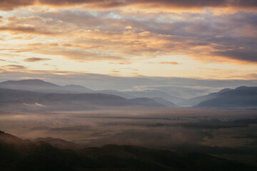 Scenic mountain landscape with golden low clouds above village among mountains silhouettes under dawn cloudy sky. Atmospheric alpine scenery of countryside in low clouds in sundown illuminating color.