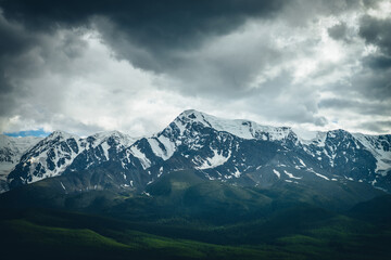 Dramatic mountains landscape with big snowy mountain ridge above sunlit forest in overcast weather. Atmospheric highland scenery with high mountain range under lead gray clouds and sunlight on forest.