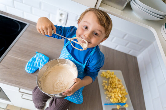 Beautiful funny little toddler boy wearing kitchen apron baking apple domestic pie. Happy child playing and working with pastry and mixer at home. Kid tasting pastry