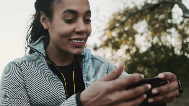 A positive young african american woman is using a phone while walking in the park outside