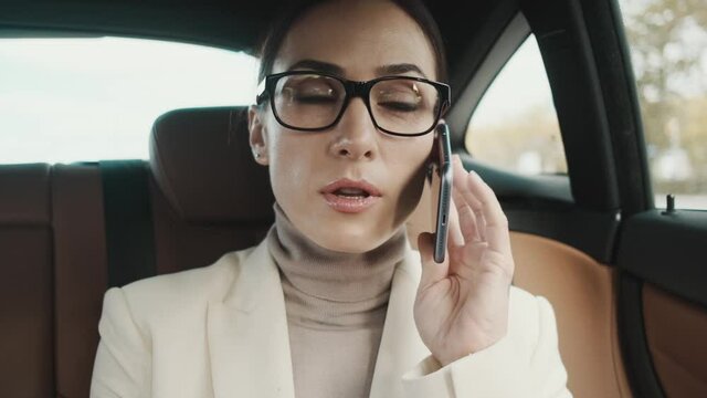 A focused young businesswoman wearing glasses is talking on her phone sitting in the car 