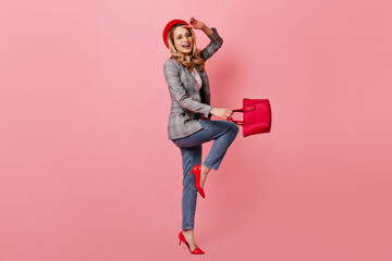 Positive girl in bright hat, jeans and jacket jumps on pink background. Full-length shot of blonde in heels with bag in her hands