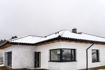 Single-family house roof covered with snow against a cloudy sky. Visible system chimney, doors, windows and falling snow.