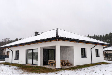 Single-family house roof covered with snow against a cloudy sky. Visible system chimney, roof trusses, windows and falling snow.