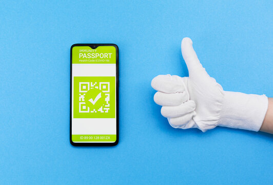 Concept of vaccination and digital passport. Hand in gloves shows thumb up and electronic Immunity passport with a COVID-19 vaccination stamp on a smartphone screen on the blue background. Flat lay