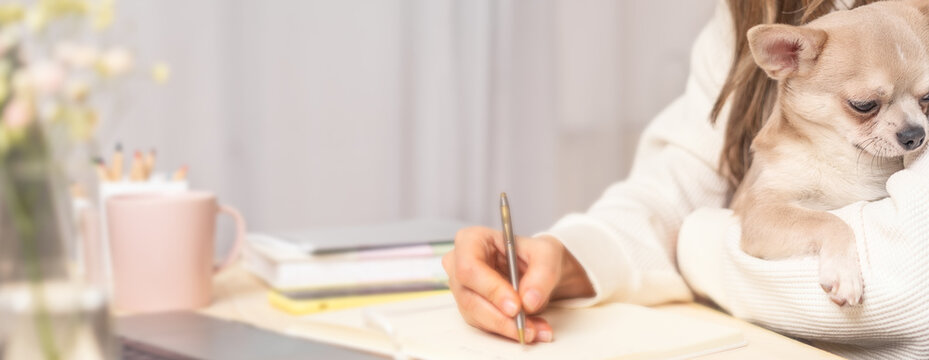 Distance learning online education and work. Business woman with puppy dog writes goals, plans, make to do and wish list in notebook on desk, working from home office. Using laptop. Focus on dog.