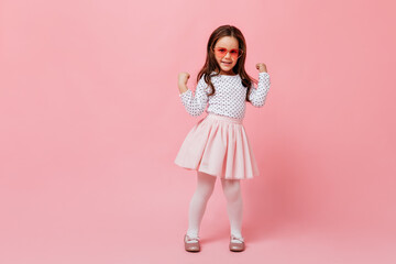 Female child in white pantyhose and dress dances against pink background. Girl in pink glasses is smiling