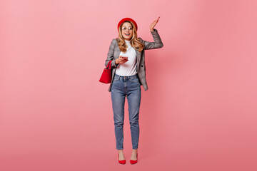 Curly blond woman with glasses and red beret shrugs. Girl in jeans and plaid jacket posing with small bag and telephone in pink studio