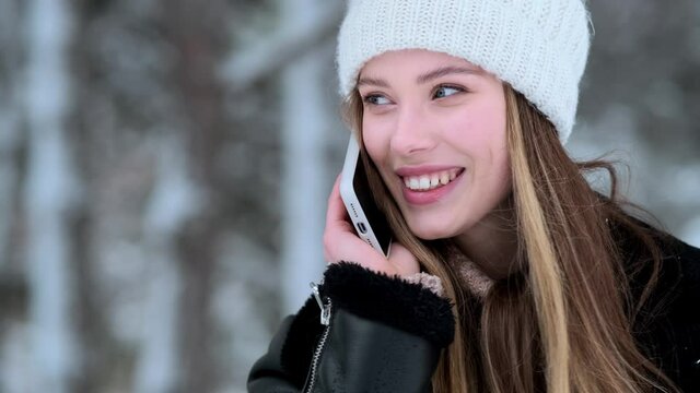 A close-up view of a beautiful young woman wearing winter hat is talking on her smartphone walking outside at wintertime