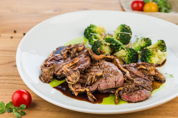 Roast beef with mushrooms and broccoli in a plate on a light wooden background.