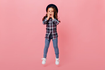 Little girl opened her mouth in surprise. Female child in jeans, blouse and black beret jumps on pink background
